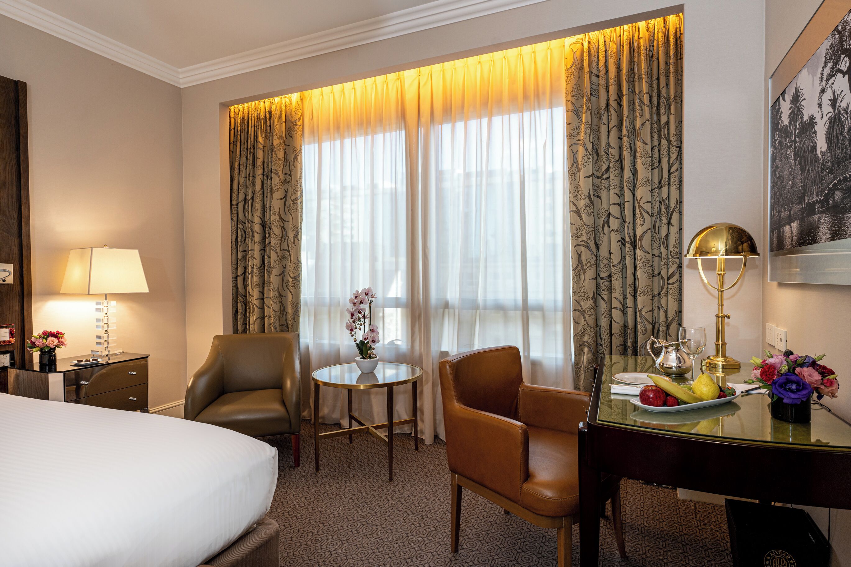 A hotel room with a bed, two leather chairs, a small round table with a lamp, a vase of flowers, and a fruit plate.