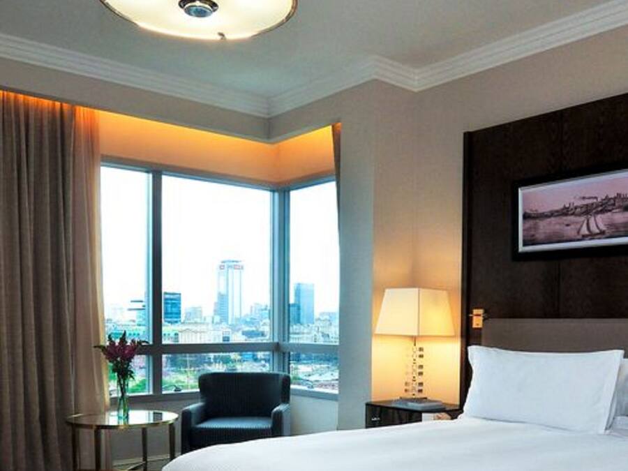 A hotel room with a large bed, a desk, a chair, a lamp, and a window with a city view.