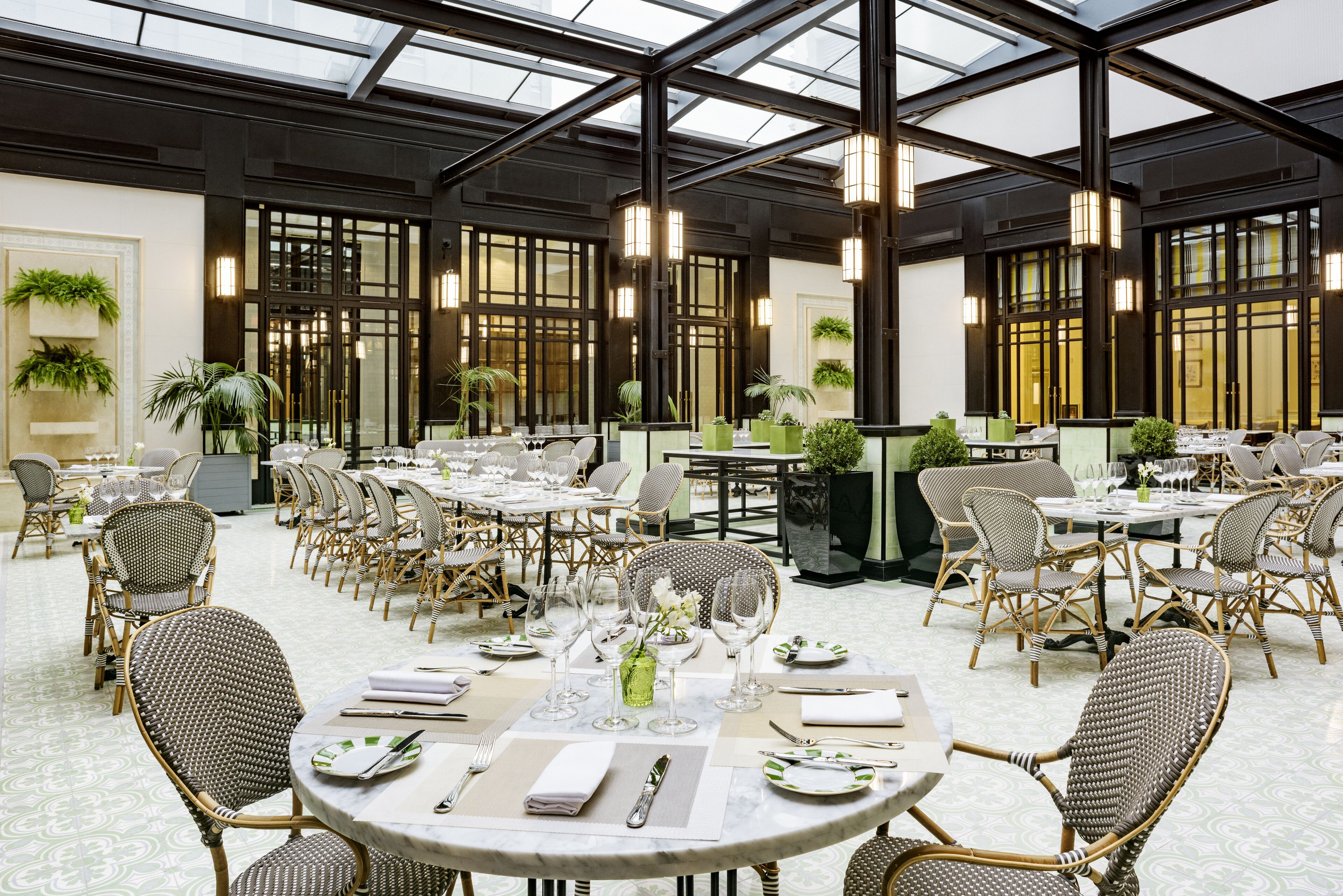 Ruime eetruimte met meerdere ronde tafels en rieten stoelen, met een hoge plafond en dakramen en grote ramen.