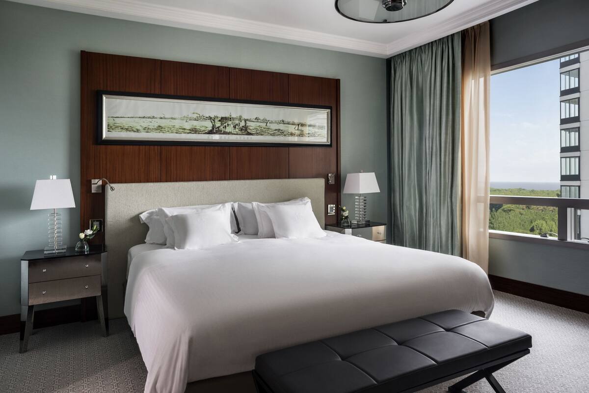 Une chambre d’hôtel avec un grand lit, deux tables de chevet avec des lampes, un tableau encadré au-dessus du lit, un banc au pied du lit et une vue sur un immeuble par la fenêtre.