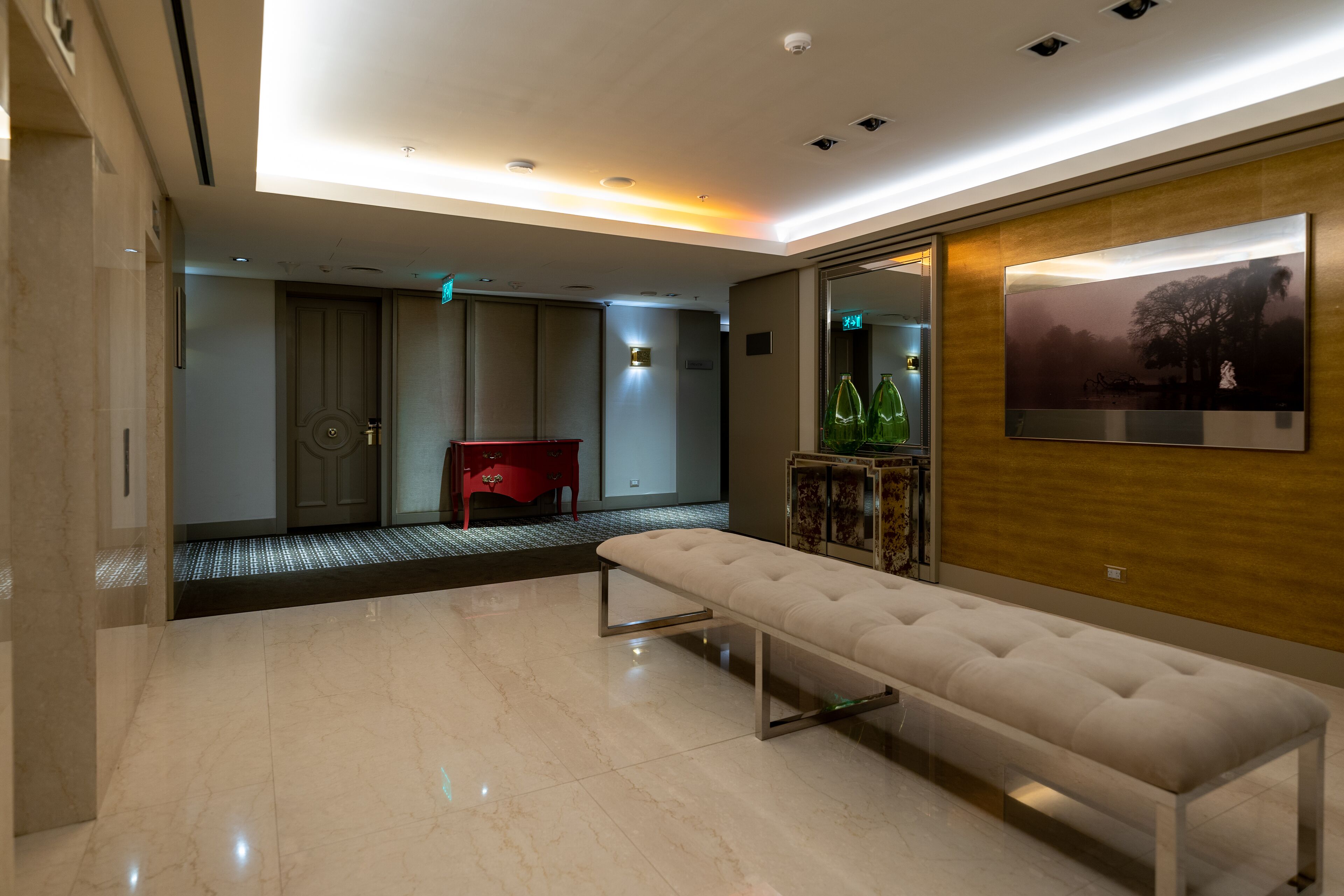 A modern hotel lobby with a marble floor, a bench, a red chair, and a wall-mounted TV.