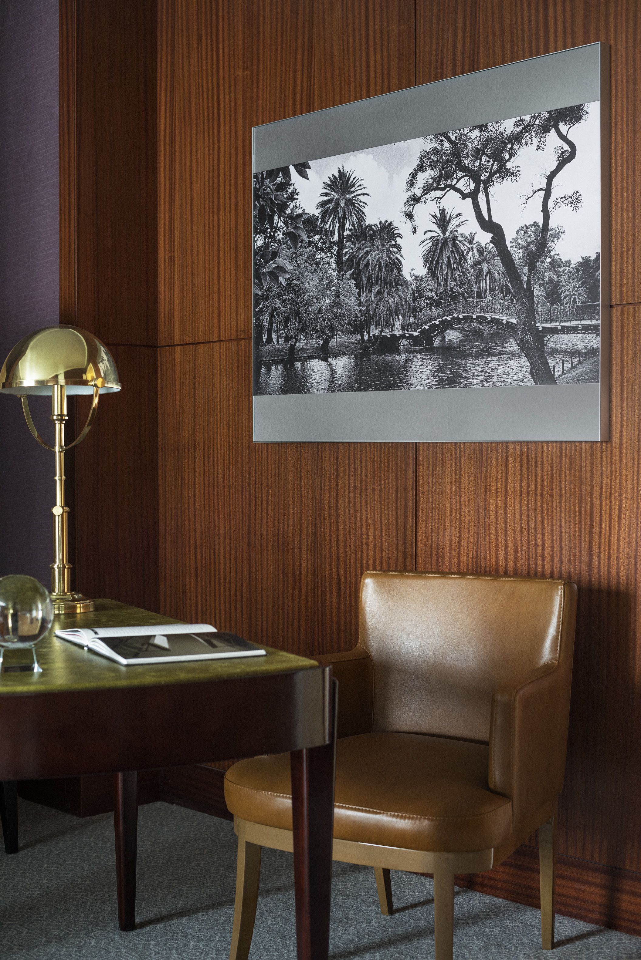 Una habitación con un panel de madera, un escritorio con una lámpara, una silla y una fotografía en blanco y negro de un puente y árboles.