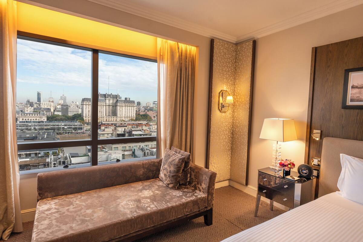 Une chambre d’hôtel avec une grande fenêtre, un canapé, une table de chevet avec une lampe et un lit.