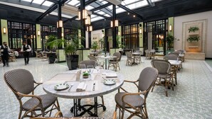 A spacious indoor area with round tables and wicker chairs, featuring a high ceiling with skylights and potted plants.