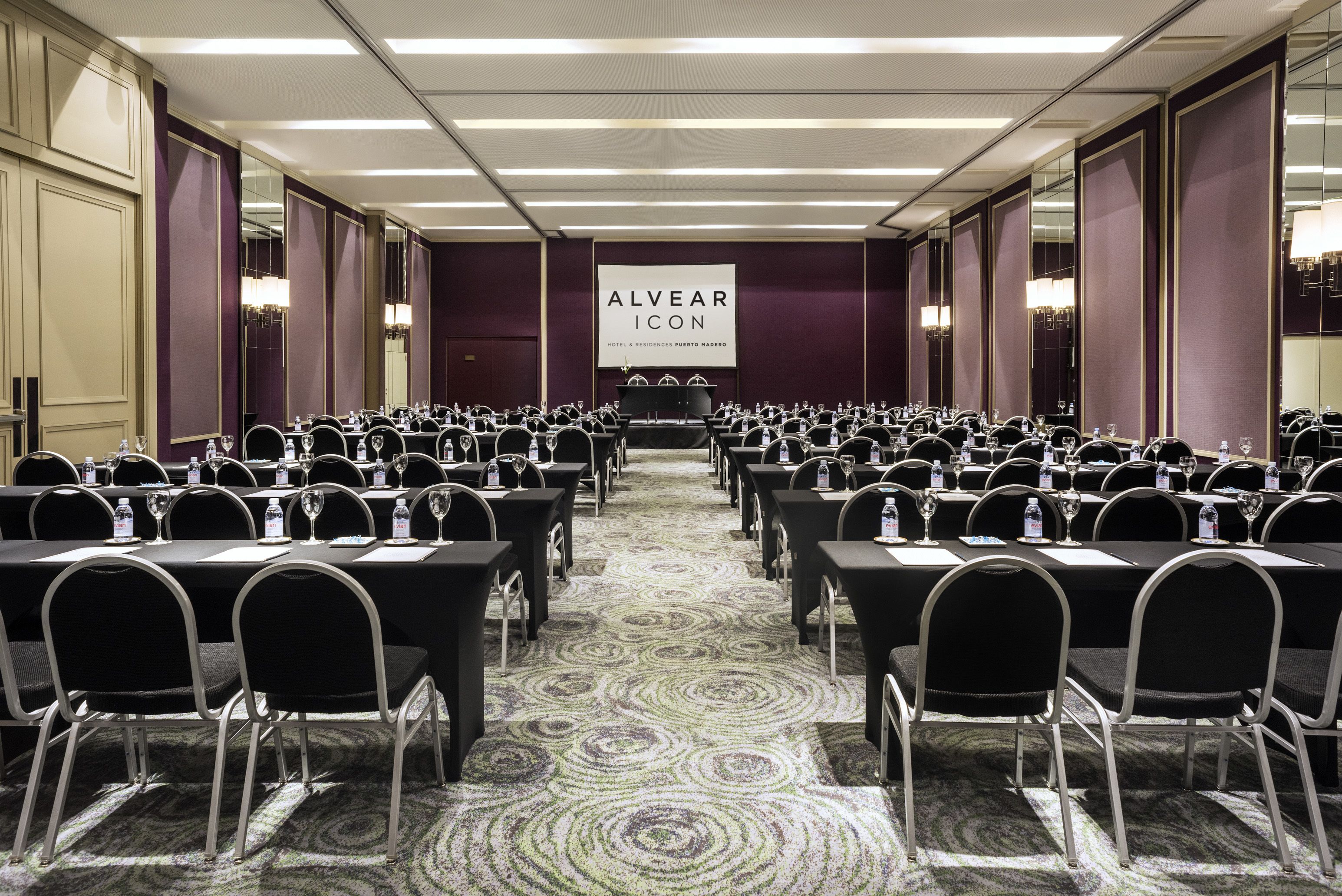Un salón de conferencias con filas de mesas y sillas, un gran cartel que dice "ALVEAR ICON HOTEL & RESIDENCES PUERTO RICO", y suelo alfombrado.