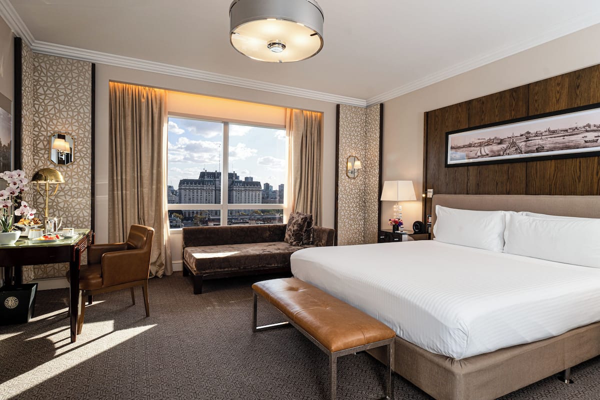Une chambre d’hôtel avec un grand lit, un bureau avec une chaise, un banc, une fenêtre avec des rideaux et une vue sur la ville.