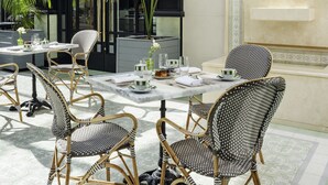 An outdoor seating area with wicker chairs and tables, surrounded by large windows and potted plants.