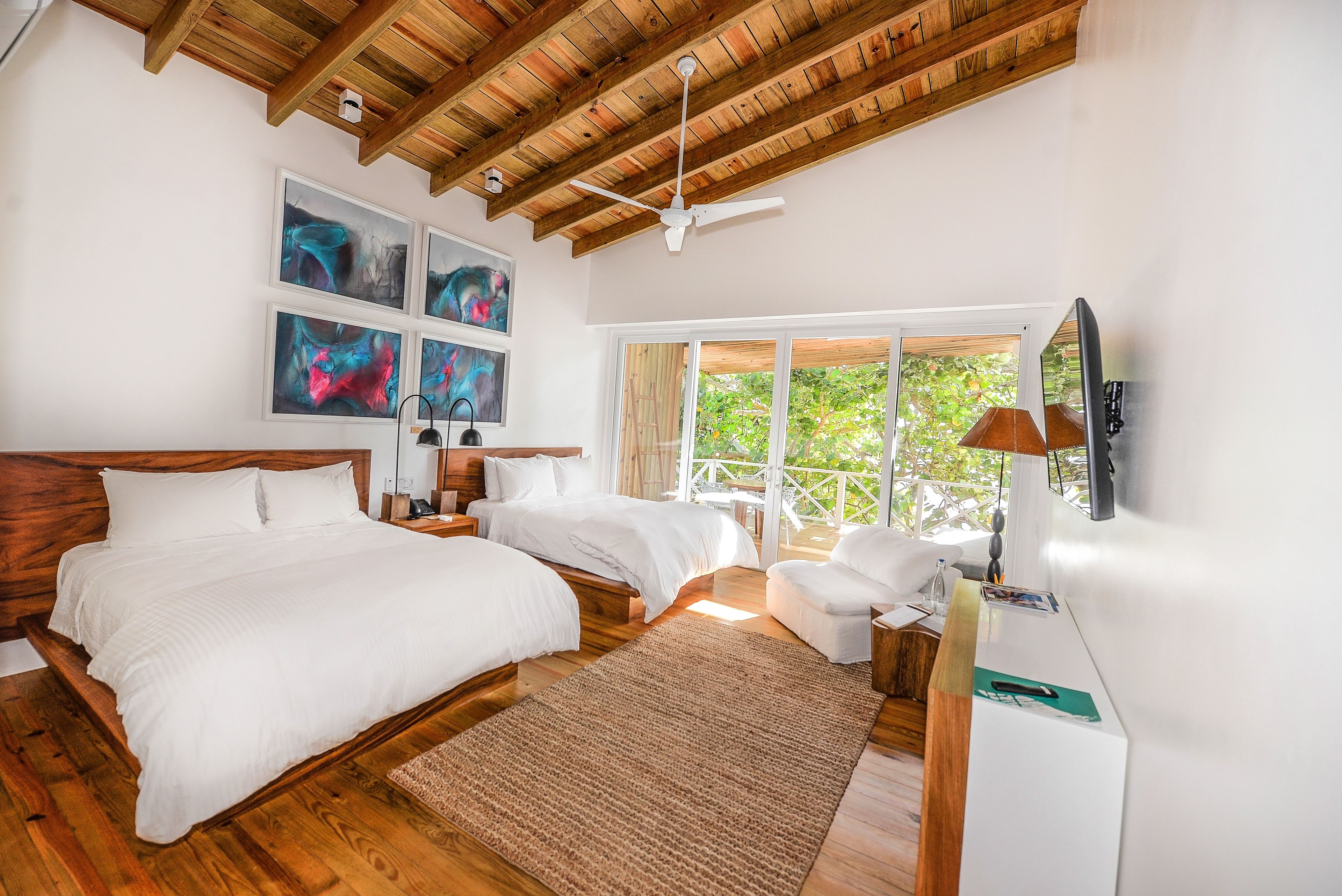 A bedroom with two beds, wooden flooring, ceiling fans, artwork, a large window, and a view of greenery.