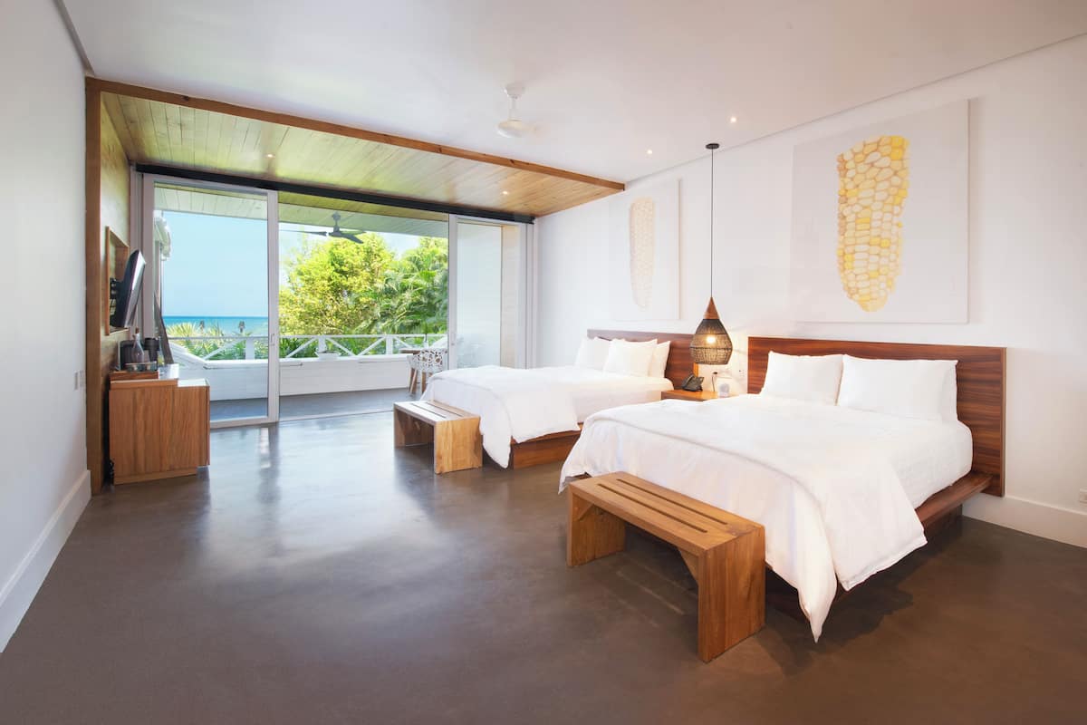 Ein modernes Schlafzimmer mit zwei Betten, Holzbanken, einem großen Fenster mit Meerblick und einem Deckenventilator.