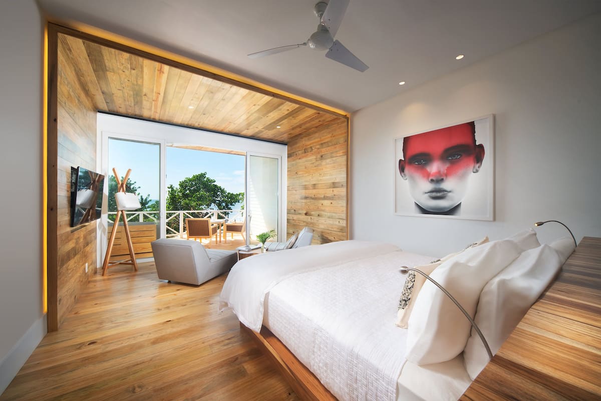 Ein modernes Schlafzimmer mit einem großen Bett, Holzboden, Deckenventilator und Blick ins Freie.