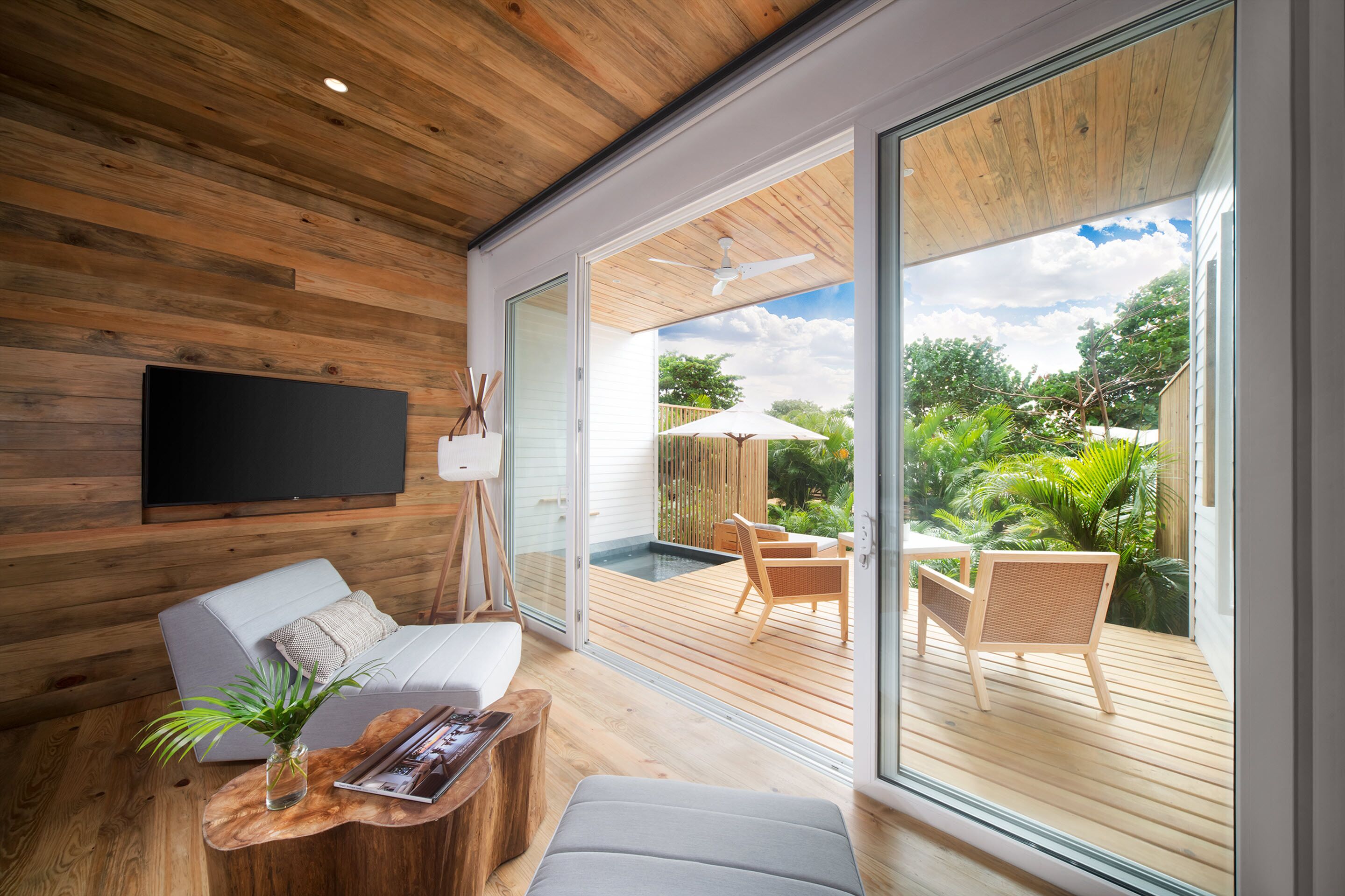 A modern living room with a wooden ceiling, a flat-screen TV, a sofa, a coffee table, and a view of an outdoor deck with a pool and chairs.