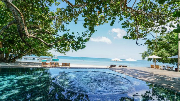 Una piscina cristalina con un jacuzzi circular, rodeada de tumbonas y sombrillas, con vistas a una playa.