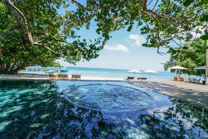 A clear pool with a circular hot tub, surrounded by lounge chairs and umbrellas, overlooking a beach.
