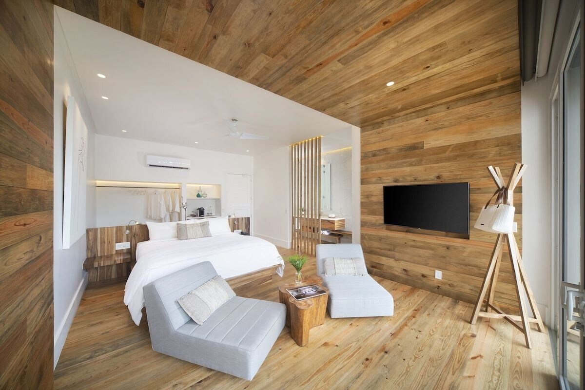 Quarto moderno com uma cama grande, duas poltronas, uma TV e piso de madeira.