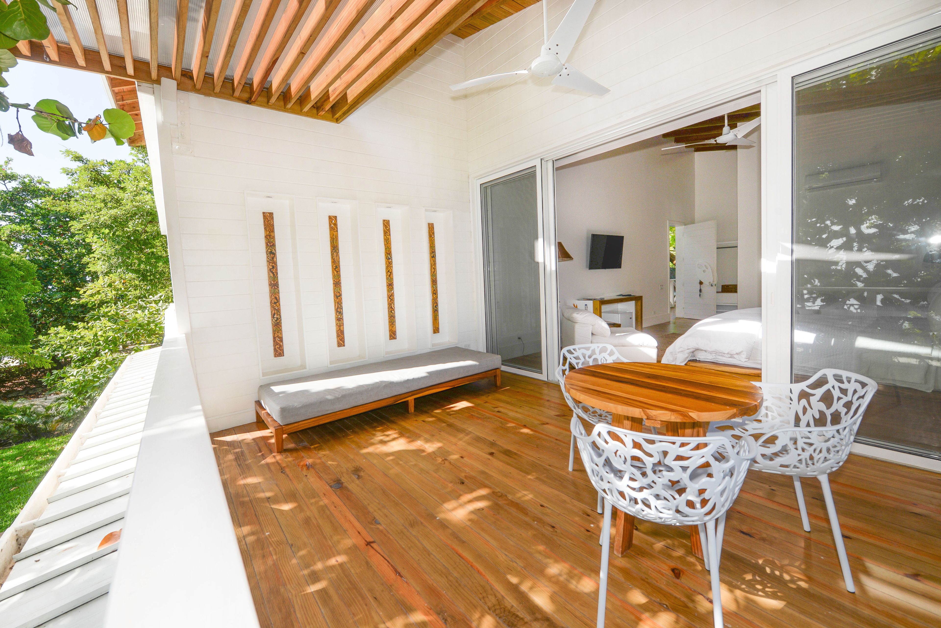 A wooden deck with a bench and two white chairs, a ceiling fan, and a sliding glass door leading to a room with a bed and a TV.