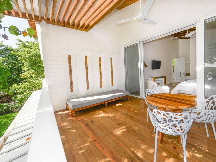 Una terraza de madera con un banco y dos sillas blancas, un ventilador de techo y un gran ventanal corredera que da a una habitación con una cama y un televisor.