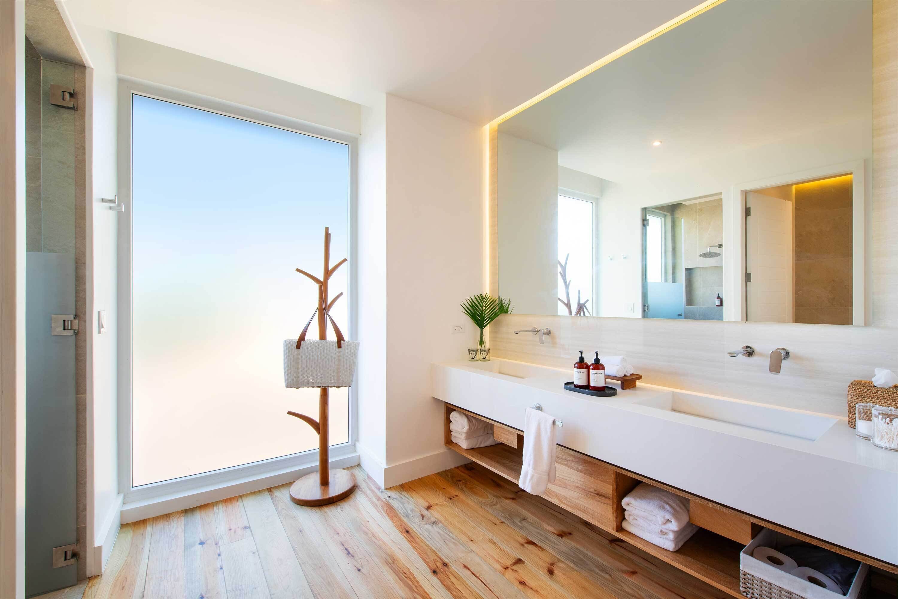 A modern bathroom with a large mirror, wooden flooring, and a view through a sliding door.