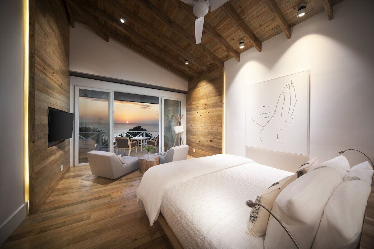 Ein modernes Schlafzimmer mit einem großen Bett, einem Flachbildfernseher, einer Holzbalkendecke und Blick auf den Sonnenuntergang durch eine Schiebetür.
