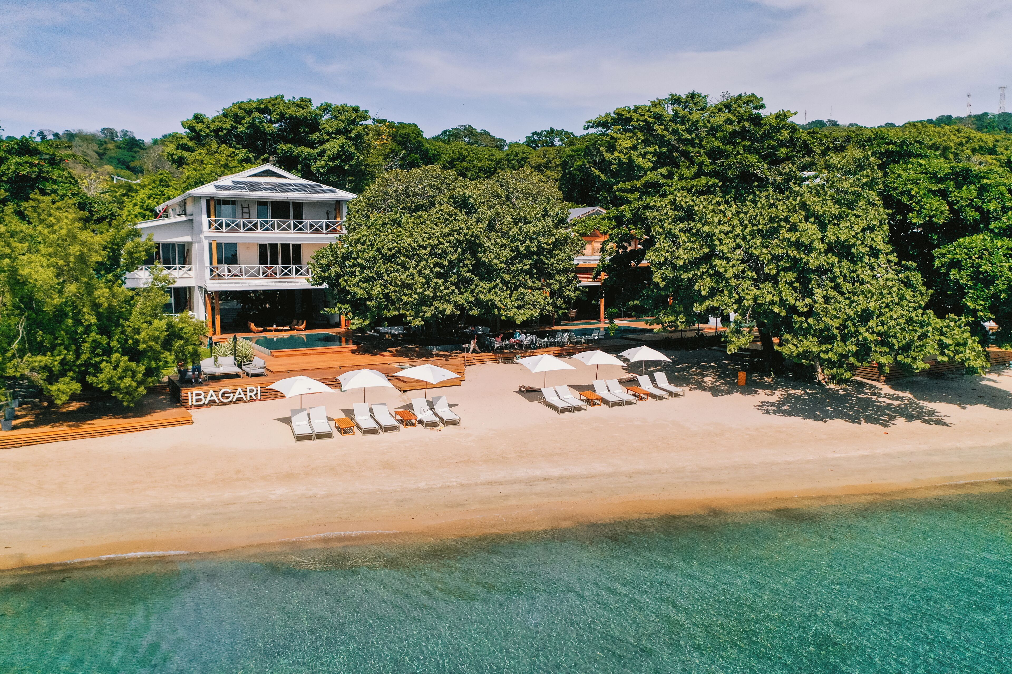 A beachfront property with a two-story building, a sandy beach with sun loungers and umbrellas, and lush greenery.