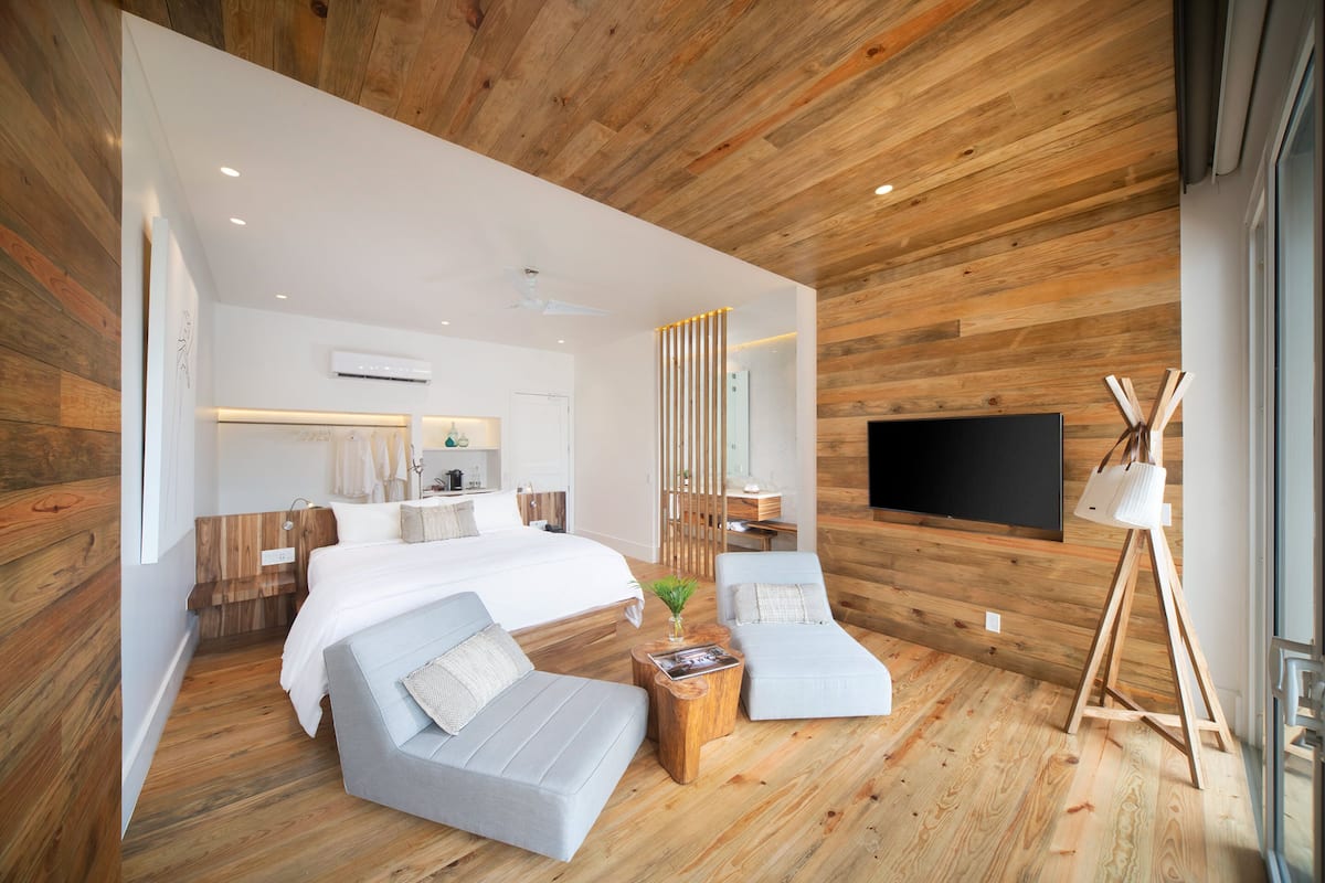 Ein modernes Schlafzimmer mit Holzboden, einem großen Bett, zwei grauen Sofas, einem Holztisch und einem an der Wand montierten Fernseher.