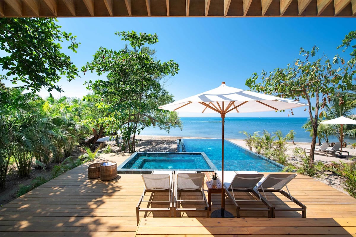 Eine Holzterrasse mit Pool, Liegestühlen und Sonnenschirmen mit Blick auf Strand und Meer.