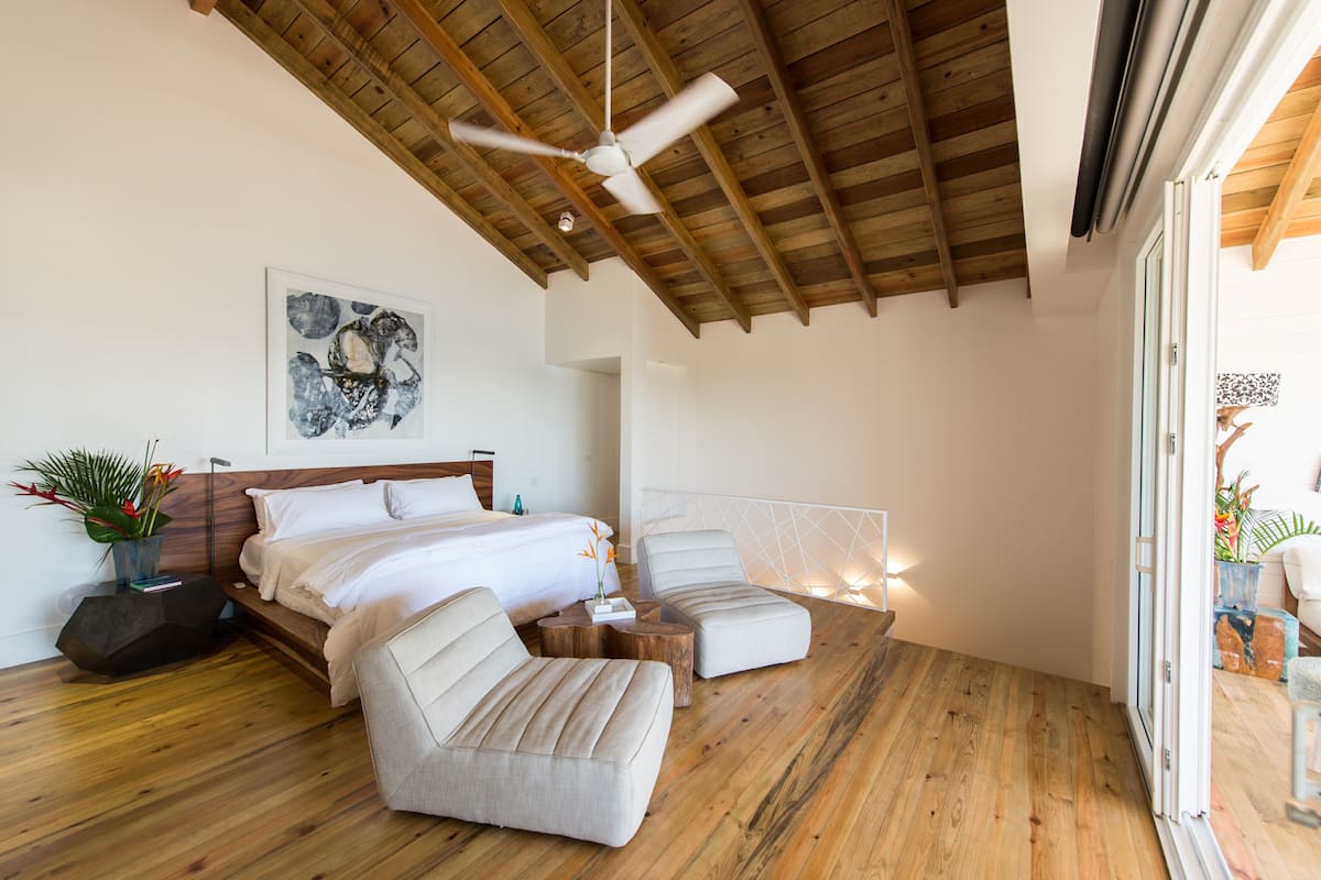 Ein modernes Schlafzimmer mit Holzboden, einem Bett, zwei weißen Sesseln und einem Nachttisch mit Blumen.