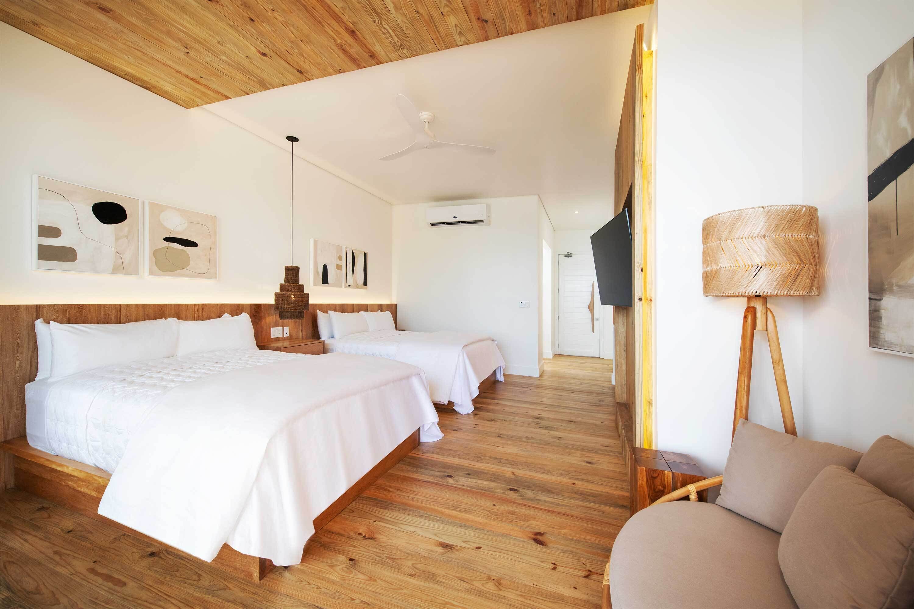 A hotel room with two beds, a wooden ceiling, a standing lamp, and abstract wall art.