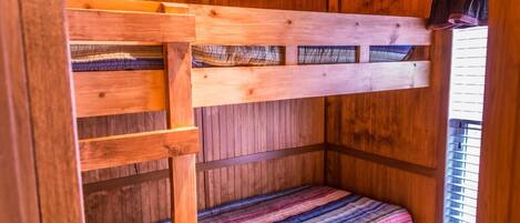 A wooden bunk bed with a striped blanket, built-in shelves, and a window with blinds.