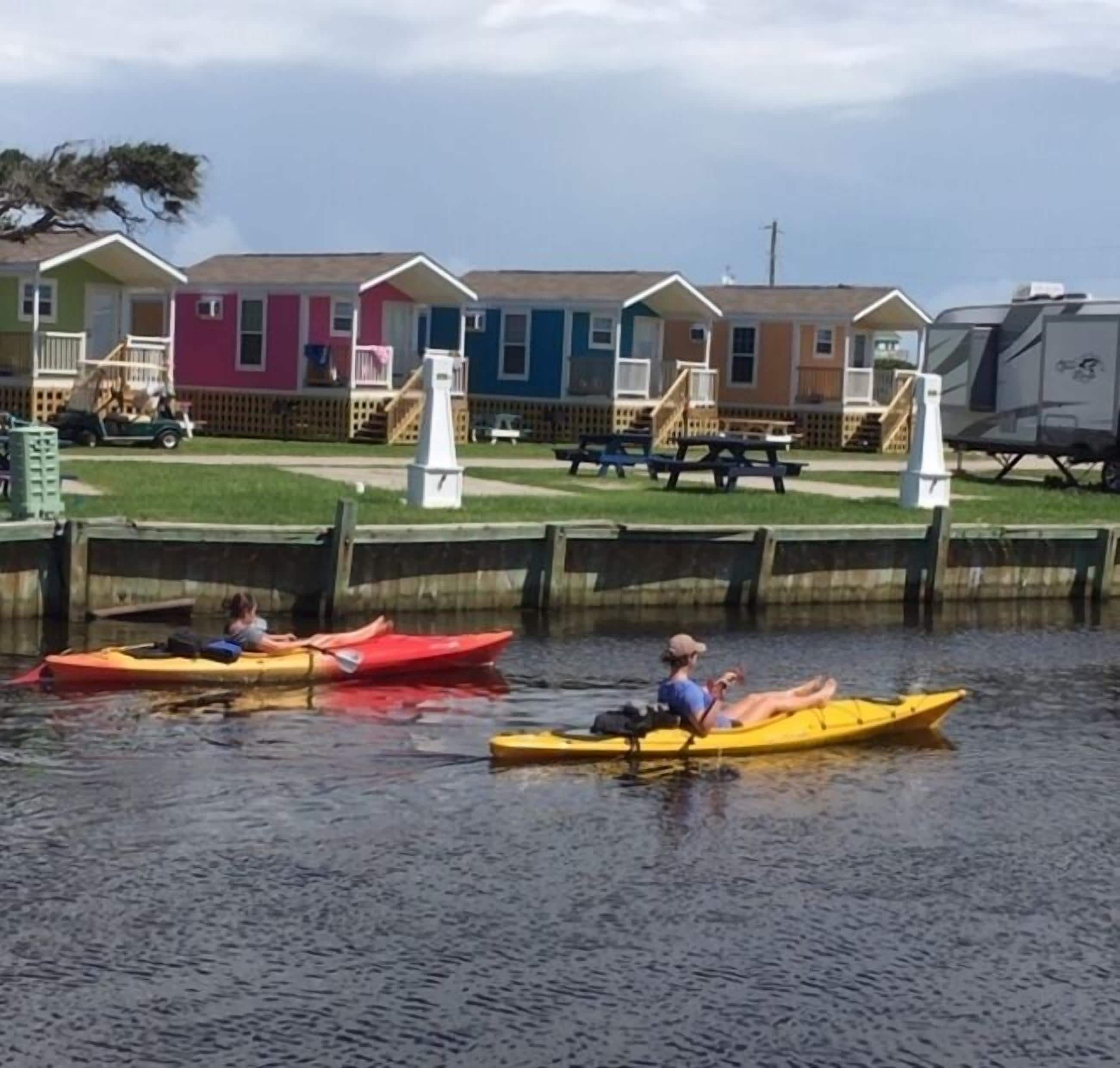Une rangée de maisons mobiles aux couleurs vives, situées au bord d’un plan d’eau paisible, avec des kayaks à proximité.