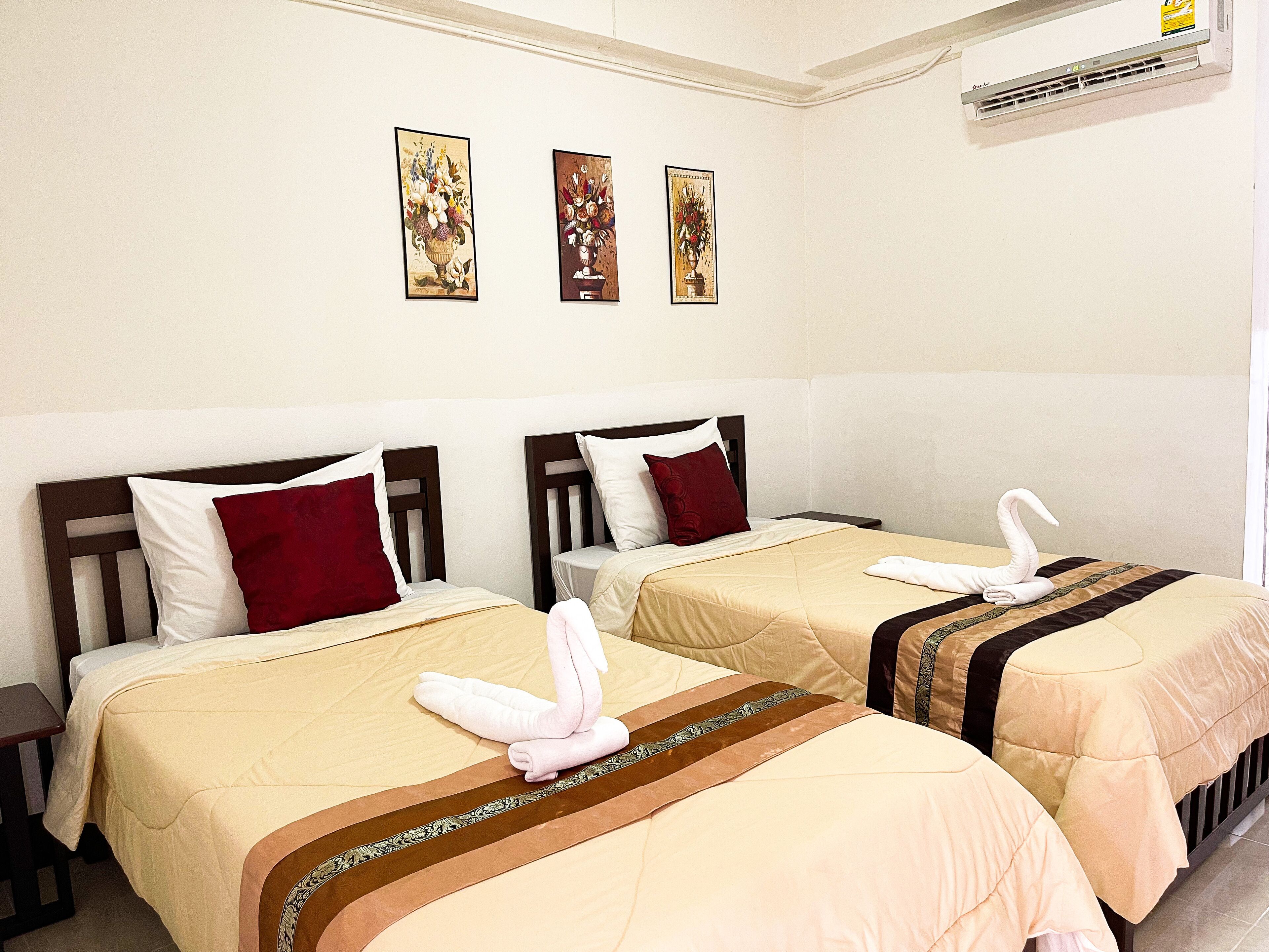 A hotel room with two beds, each with a red and white pillow, beige bedding, and a decorative towel on the bed. There are three wall-mounted paintings above the beds.