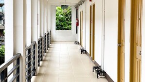 A long, narrow corridor with white walls and doors on one side, leading to a green area outside.