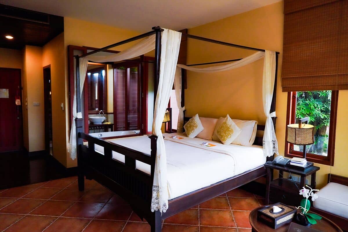 A four-poster bed with white linens and gold pillows, a bedside table with a lamp, and a window with a view of greenery.