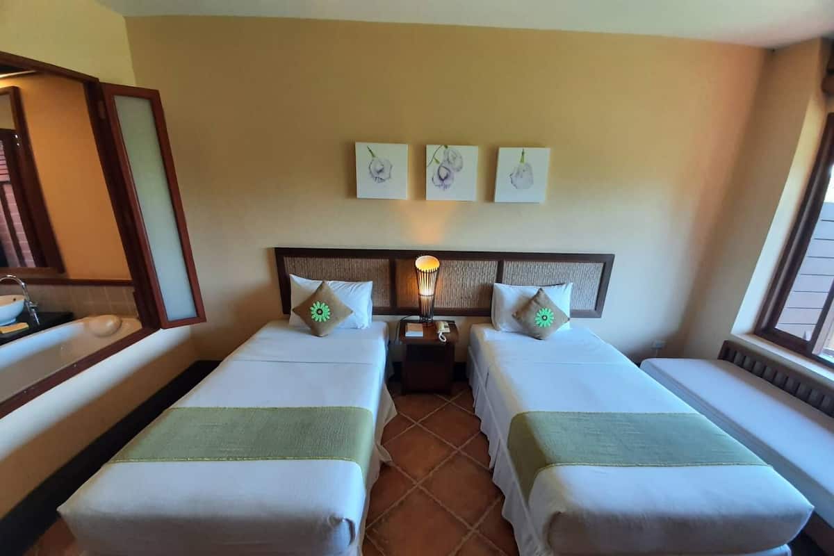 A hotel room with two beds, a nightstand with a lamp, a bathroom with a sink and mirror, and a window with blinds.