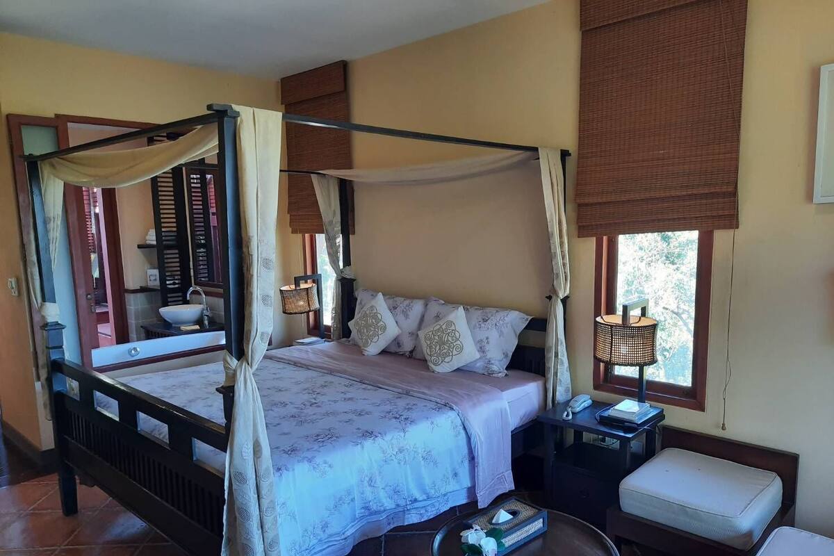 A four-poster bed with a canopy, a bedside table with a lamp, a small round table with a vase, and a bench.