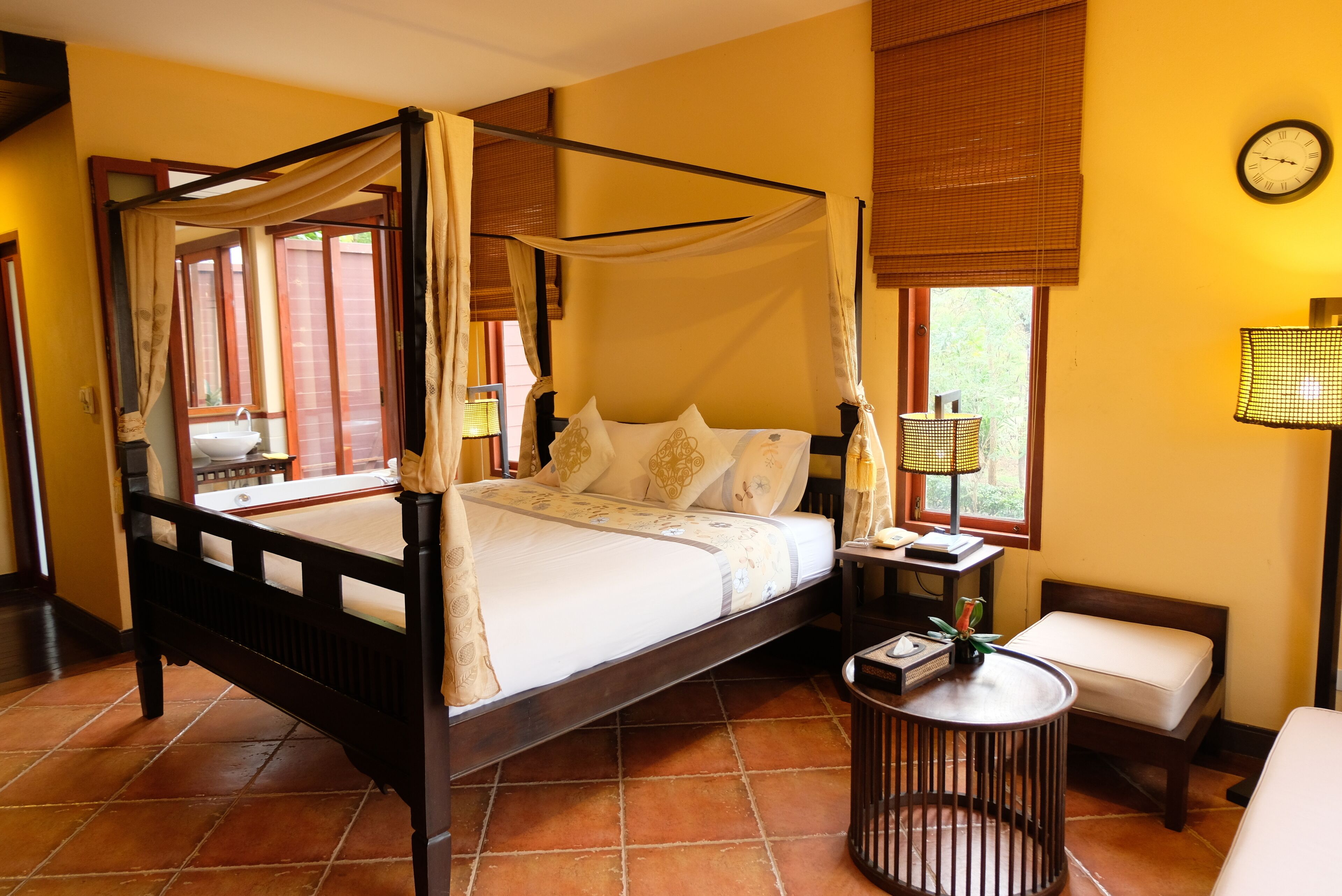A four-poster bed with a canopy, a bedside table with a lamp, a small bench, and a clock on the wall.