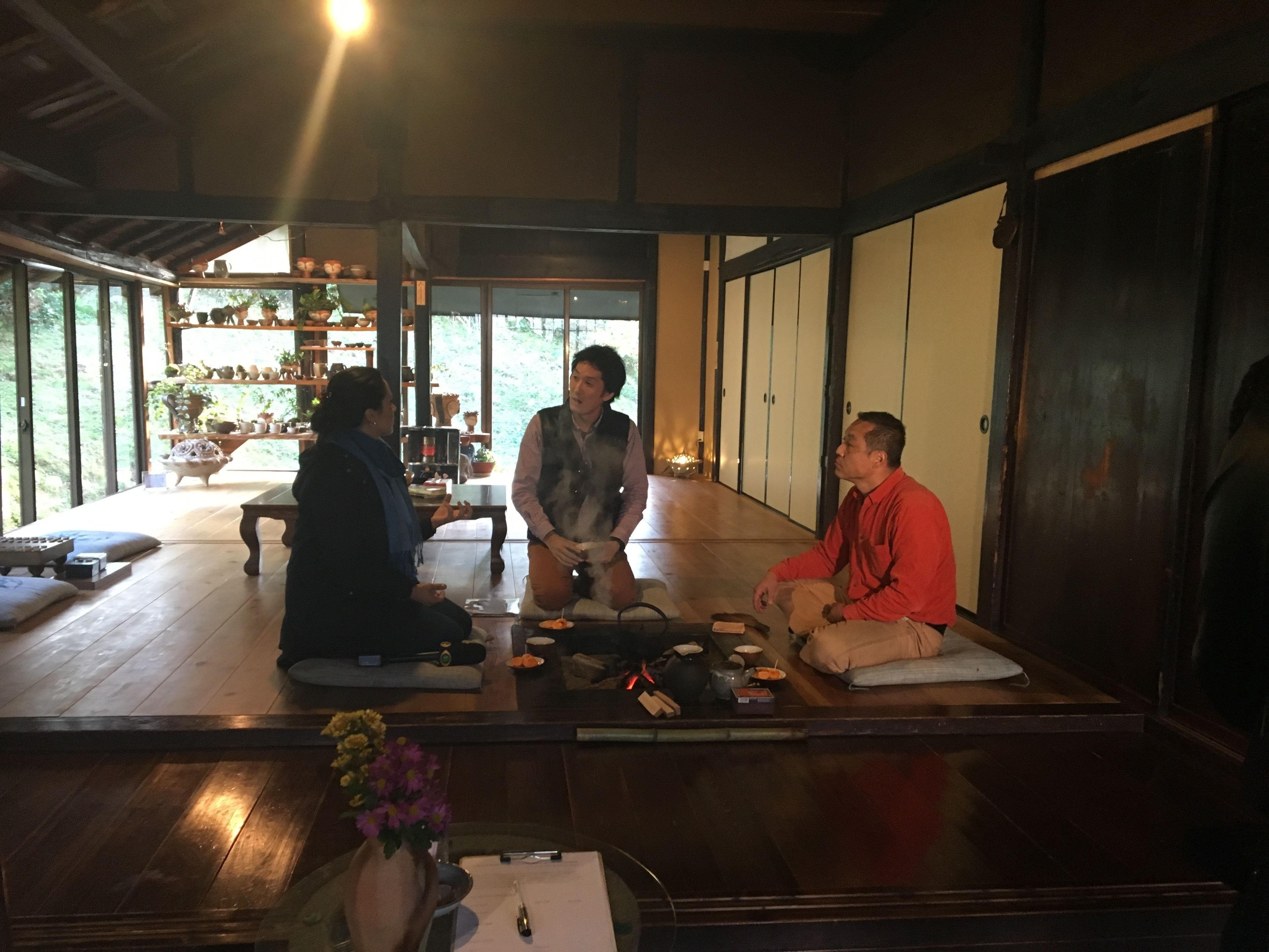 A traditional Japanese room with tatami flooring, a low table with a vase of flowers, and a wooden shelf with various items.