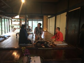 Una habitación japonesa tradicional con suelo de tatami, una mesa baja con un jarrón de flores y una estantería de madera con diversos objetos.