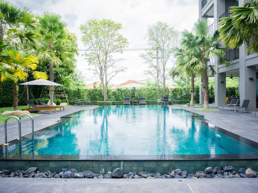 A swimming pool with lounge chairs and a covered seating area, surrounded by palm trees and a building with balconies.