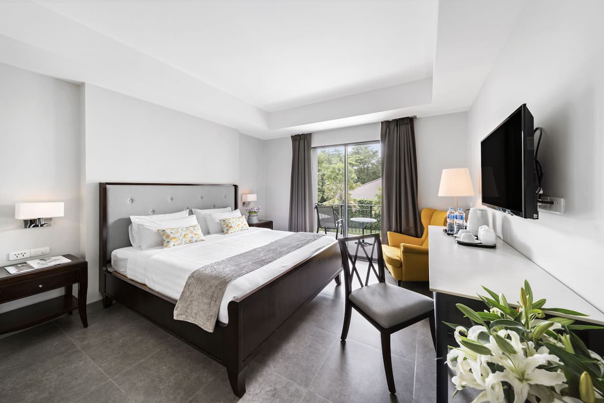 A modern hotel room with a large bed, a desk, a chair, a TV, and a balcony with a view of greenery.