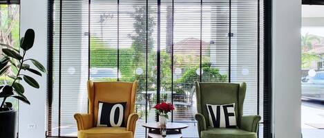 A modern room with two armchairs facing a large window with blinds, a small round table with a vase of flowers, and a potted plant to the left.