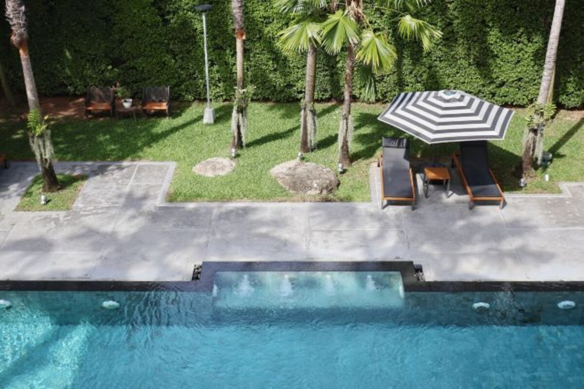 Una piscina con zona de descanso, que incluye sillas y una sombrilla, rodeada por un jardín ajardinado con palmeras y setos.