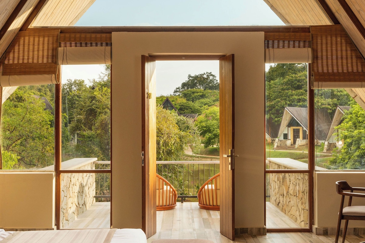 A bedroom with a large bed, a bench, and a view of a balcony with a stone wall and greenery.