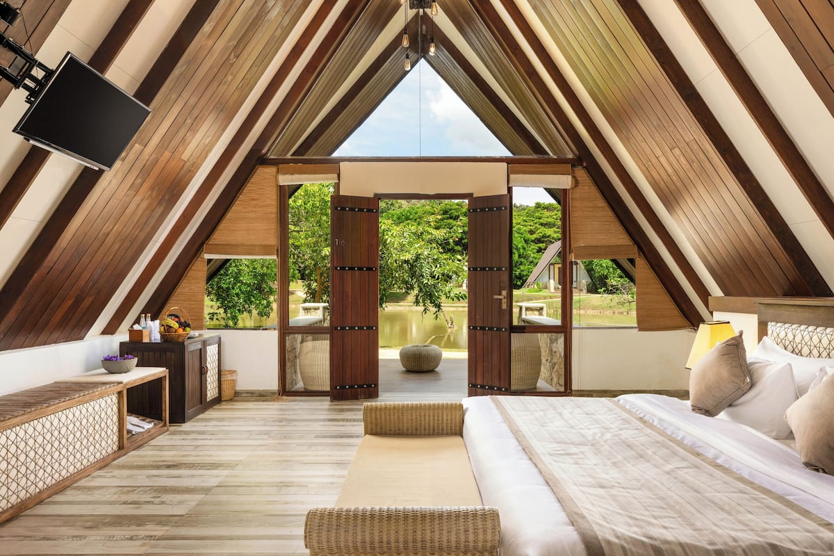 A high-ceilinged room with a bed, a TV, and a view of greenery outside.