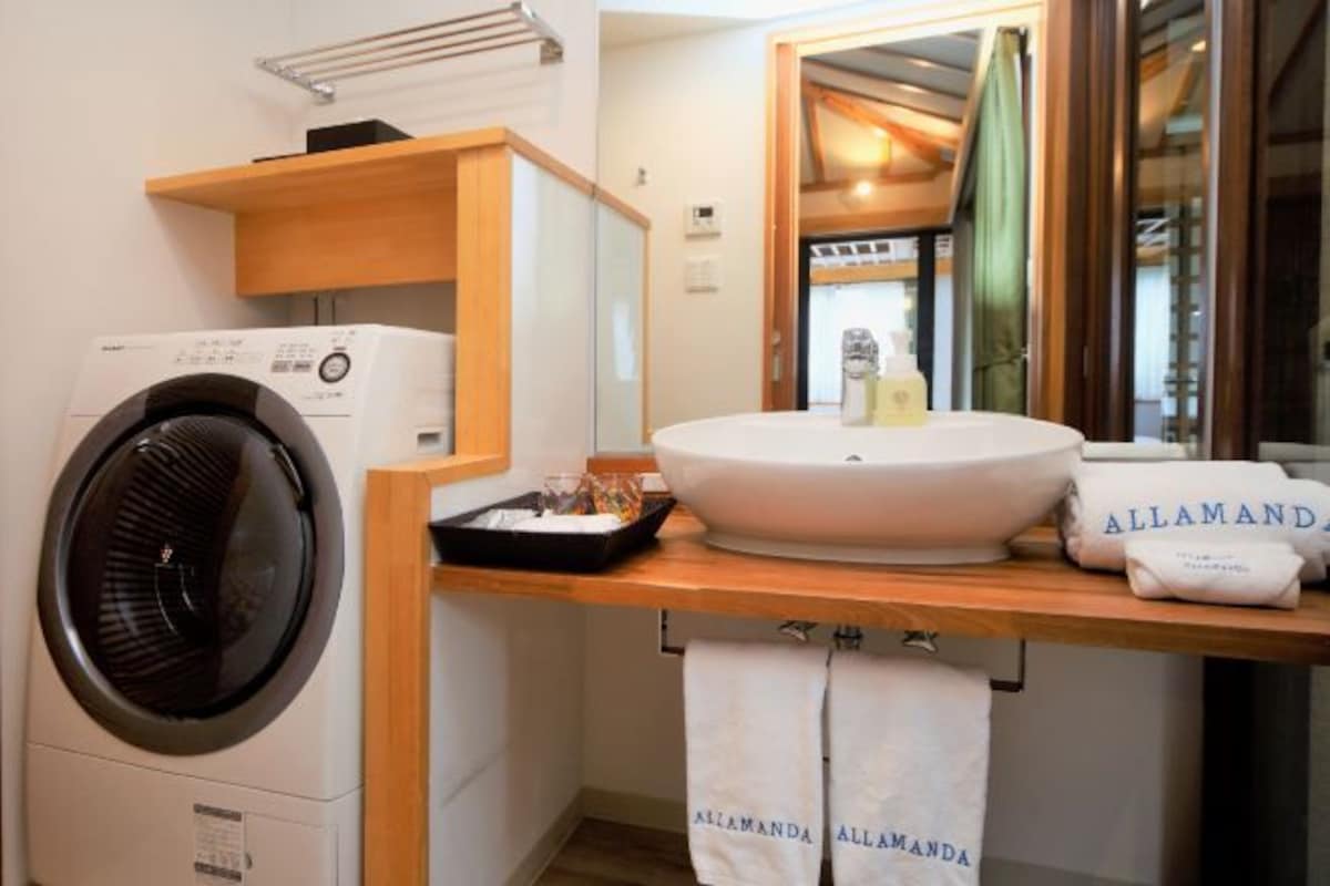 Een hotelbadkamer met een wasmachine, een wastafel en handdoeken met de merknaam 'ALLAMANDA'.