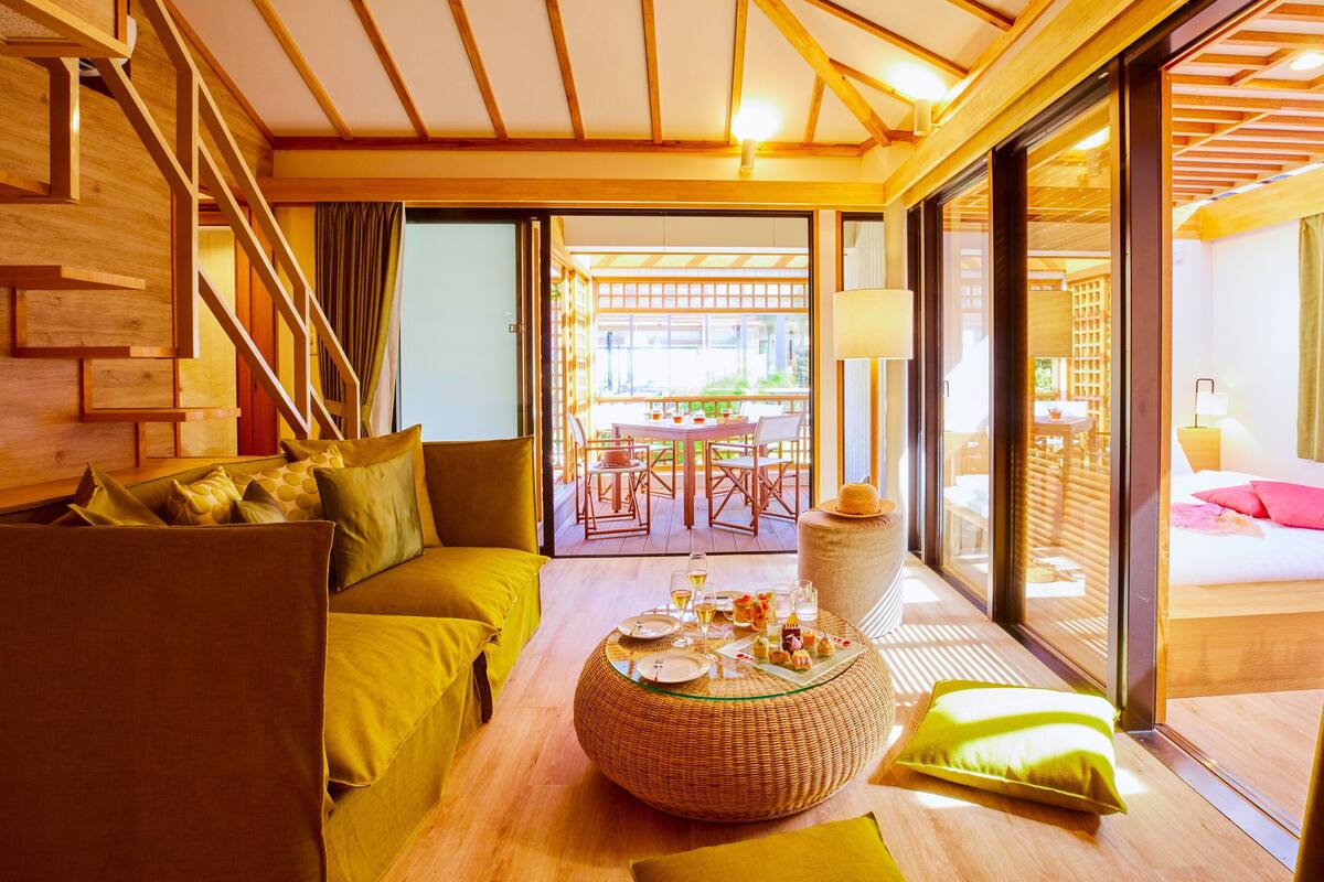 Ein modernes Interieur mit Holzboden, einer Sofagarnitur, einem Esstisch mit Korbschmuck und Blick durch eine Glastür auf einen Außenbereich.