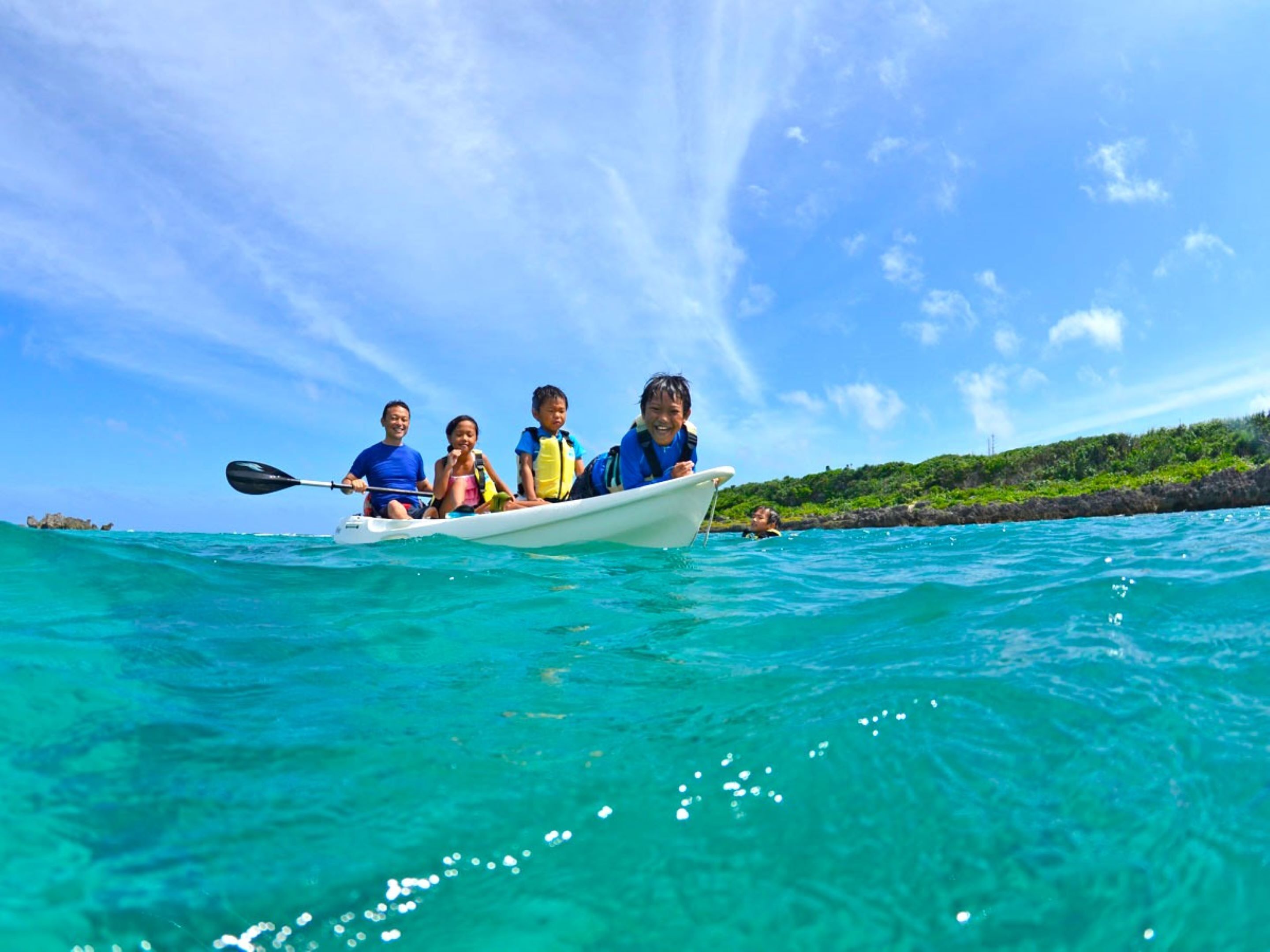 A group of people in a small boat on turquoise water, with a clear sky and a rocky coastline in the background.