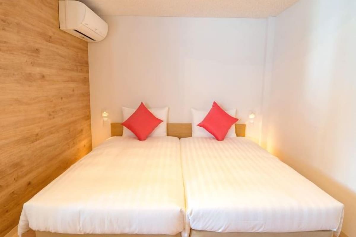 Cama doble con ropa de cama blanca y almohadas rojas, pared de madera y equipo de aire acondicionado en el techo.