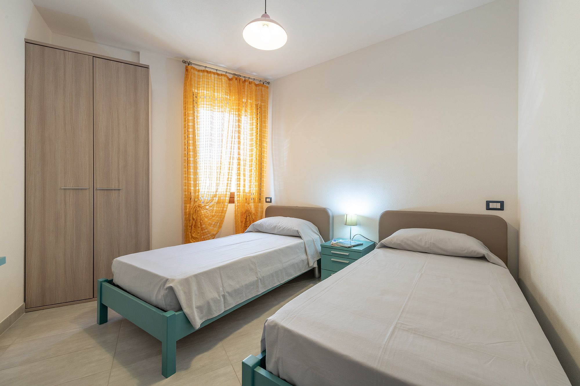 Un dormitorio con dos camas individuales, un armario, una mesita de noche con lámpara y una ventana con cortinas amarillas.