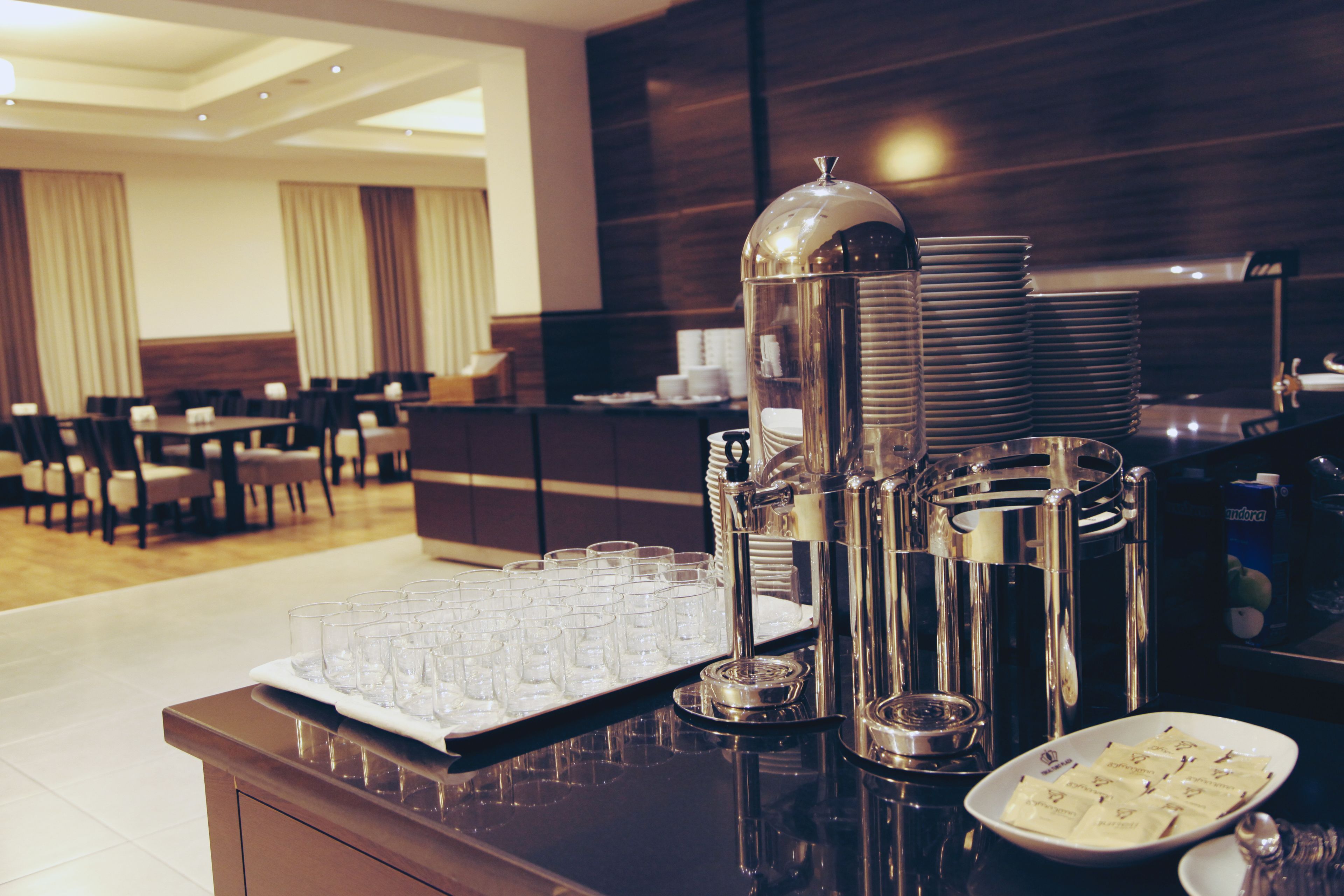 Zona de recepción de hotel con una barra de autoservicio que incluye una bandeja de vasos, un dispensador de café y un plato con paquetes de comida.