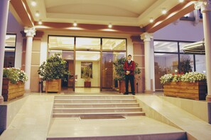 Ein Hotel Eingang mit einem Mann in Uniform, der vor den Stufen steht. Auf jeder Seite des Eingangs befinden sich Kübelpflanzen.
